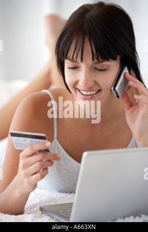 Junge Frau auf Bett liegend mit Laptop, Kreditkarte zu halten, während Mobiltelefon, Porträt Stockfoto