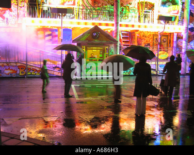 fairground ride with box office and spectators with umbrellas reflected in wet pavement Stockfoto