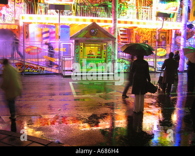 fairground ride with box office and spectators with umbrellas reflected in wet pavement Stockfoto