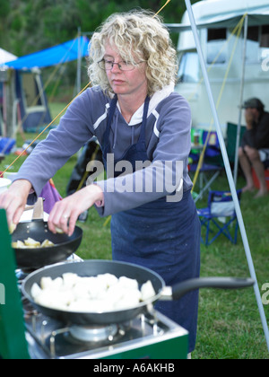 Frau, die Zubereitung von Speisen in einem externen Lager Ambiente mit Gas gefeuert Ofen bereitet Essen mit Zelten und Stühle im Hintergrund Stockfoto