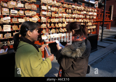 Zwei Frauen hängen eine hölzerne Tafel mit Wünsche darauf geschrieben, außerhalb des wichtigsten Tempels im Hachimangu Schrein in Kamakura, Japan Stockfoto