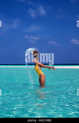 Hübsche junge blonde Frau im gelben Badeanzug stehen in der Lagune der Malediven im Indischen Ozean kippen Wasser aus ihren langen Haaren Stockfoto