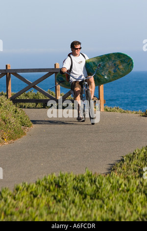 Ein Mann, mit dem Fahrrad mit einem Surfbrett über dem Ozean in Santa Cruz, Kalifornien Stockfoto