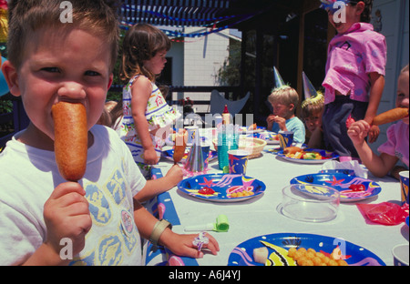 Porträt eines jungen Mannes, einen Mais-Hund zu essen, während die anderen Kinder auf einer Geburtstagsfeier im Hintergrund spielen Stockfoto