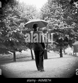 Mann mit Bowler Hut mit Regenschirm und Aktentasche, der draußen im Regen steht. Schwarz-weiß. Stockfoto