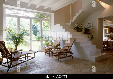 Rattan Chaiselongues Terrakottafliesen Stock in Halle mit weiße Treppe und Blick auf Garten durch Glastüren Stockfoto