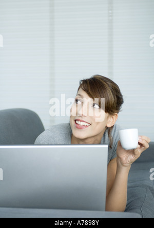 Junge Frau auf Couch, mit Laptop und halten Kaffee Tasse, nachschlagen Stockfoto