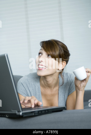 Junge Frau auf Couch, mit Laptop und halten Kaffee Tasse, nachschlagen Stockfoto