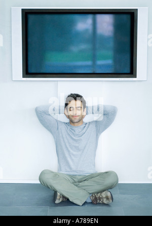 Mann sitzt auf dem Boden unter Breitbild-TV, Hände hinter den Kopf und die Augen geschlossen Stockfoto