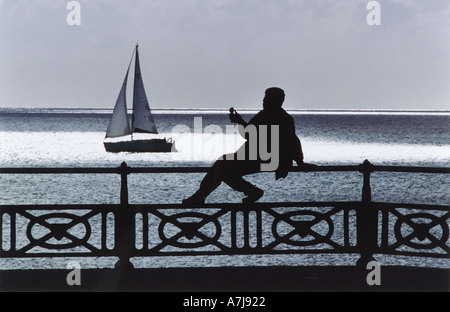 Strandpromenade der Stadt Brighton und Hove. Eine rundliche Frau sitzt am Geländer, Eis essen, wie eine Yacht vorbei fährt. Stockfoto
