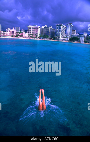 Eine Frau, Eintauchen in den kristallklaren Gewässern vor Waikiki Hawaii unter einem bewölkten Himmel mit der Skyline im Hintergrund Stockfoto