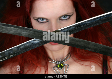 Frau mit roten Haaren eine Spinne Halskette trägt und hält zwei gekreuzte Schwerter vor ihr Gesicht Stockfoto