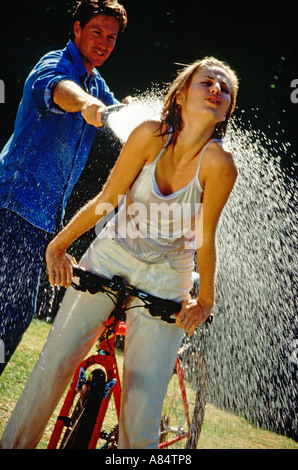 Junges Paar im Freien im Garten.  Mann Spritzen Wasser auf Frau Reiten Fahrrad. Stockfoto