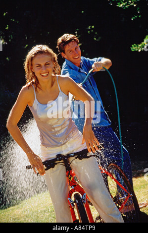 Junges Paar im Freien im Garten.  Mann Spritzen Wasser auf Frau Reiten Fahrrad. Stockfoto