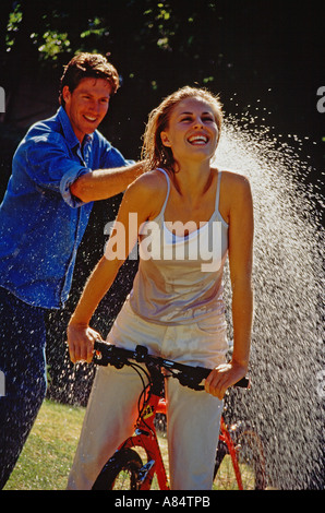 Junges Paar im Freien im Garten.  Mann Spritzen Wasser auf Frau Reiten Fahrrad. Stockfoto