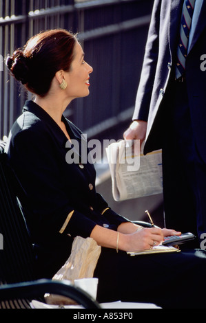 Nahaufnahme der elegant gekleidete Frau sitzt auf der Bank lächelte nicht identifizierbare Mann im Anzug stehen im Freien im urbanen Umfeld. Stockfoto