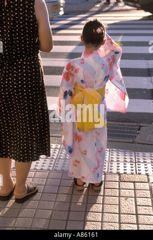 Ein kleines Mädchen, gekleidet in einem niedlichen Sommer Yukata robe mit gelbe Schärpe warten mit ihrer Mutter die Straße überqueren Stockfoto