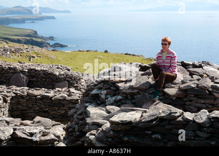 junge Frau sitzt auf einer Hütte in Form einer Bienenwabe, gebaut aus Steinen auf der Dingle-Halbinsel an der westlichen Küste von Irland Stockfoto