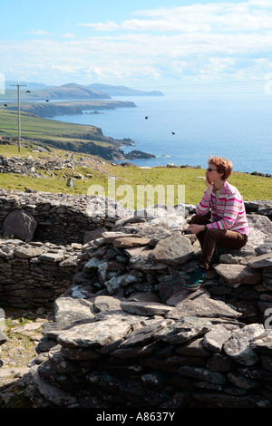 junge Frau sitzt auf einer Hütte in Form einer Bienenwabe, gebaut aus Steinen auf der Dingle-Halbinsel an der westlichen Küste von Irland Stockfoto