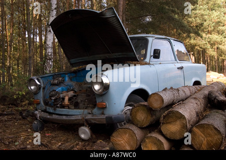 Altes, zerstörtes und verlassenes hellblaues Trabant-Auto mit offener Motorhaube in einem polnischen Wald, umgeben von gehäuften Baumstämmen und Bäumen. Stockfoto