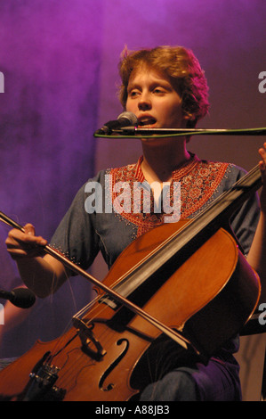 Ein Mitglied DER WARSCHAUER DORFKAPELLE tritt live im Konzert auf, spielt Cello und singt in ein Mikrofon auf einer Bühne mit atmosphärischem lila Licht. Stockfoto