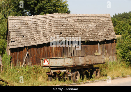 Altes Holzschuppen auf dem polnischen Land, Polen, mit einem verwitterten Fliesendach und einem rustikalen Bauernanhänger, der an einem sonnigen Tag davor geparkt wird. Stockfoto