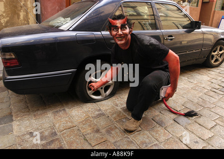 Lächelnder Teufel kniet vor einem schwarzen Mercedes-Auto mit einem Platten Reifen auf einer Kopfsteinpflasterstraße. Stockfoto