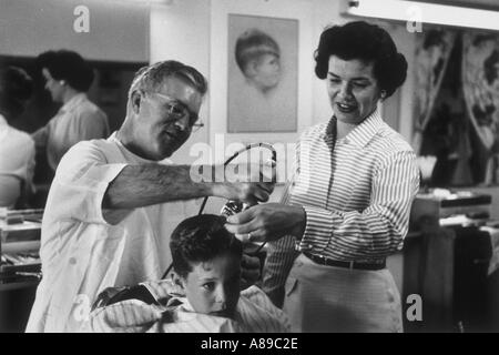 Kleiner Junge immer seinen ersten Haarschnitt von Barbier, während seine Mutter auf Bild sieht aufgenommen in den 1950 s Stockfoto