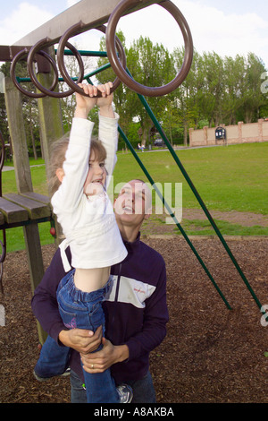 Ein kleines Kind hängt an den Affenringen eines Klettergerüsts, das von ihrem Vater in einem britischen Park hochgehoben wurde Stockfoto