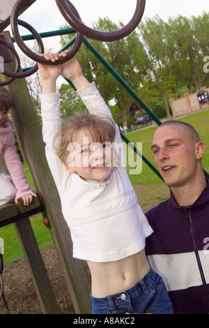 Ein kleines Kind hängt an den Affenringen eines Klettergerüsts, das von ihrem Vater in einem britischen Park hochgehoben wurde Stockfoto