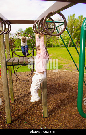 Junge kaukasische Mädchen schwingen von Metallröhrenringen auf einem Klettergerüst, mit einem weiteren Kind in der Ferne, das auf das obere Deck klettert, in einem Park Stockfoto