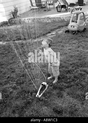 Kleiner Junge spielt mit Rasen-Sprinkler-Trinkwasser und Abkühlung an einem heißen Sommertag in seinem Hinterhof Stockfoto