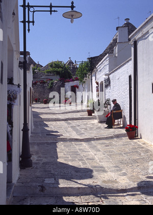 Traditionelle Trulli in einer Straße mit einem älteren Herrn in einem Stuhl im touristischen Viertel von Alberobello, Apulien, unter einem klaren blauen Himmel ruhen Stockfoto