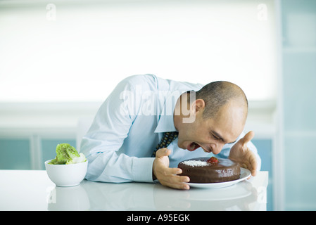Mann, sitzend am Tisch mit Schokoladenkuchen und Schüssel mit Salat, beugte sich zu Kuchen mit weit offenem Mund Stockfoto