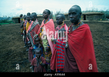 Masai Frauen und Kinder in bunten Gewändern stehen im Mittelpunkt ihres Dorfes in der Nähe ihrer Hütten Masai Mara Wildreservat Kenia Stockfoto