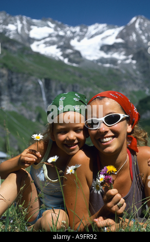 Mutter und Tochter auf alpinen Wiese liegend mit Blumen in der Hand, mit verschneiten Berglandschaft hinter. Stockfoto