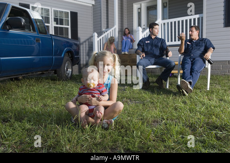 Junges Mädchen und ein Baby-sitter auf dem Rasen eines Anhängers Heim, Familie im Hintergrund Stockfoto