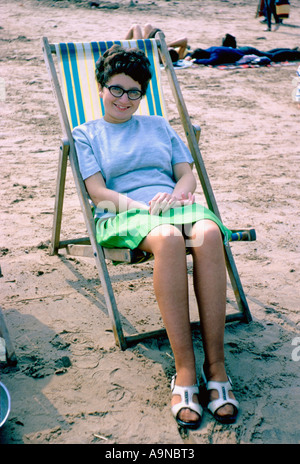 1960er Jahre: Junge kaukasische Frau in Minirock und Katzenaugenbrille, entspannend in einem Liegestuhl an einem britischen Strand Stockfoto