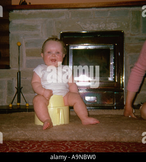 Lächelndes Baby, das auf einem Töpfchen vor dem Feuer sitzt, um die 1960er Jahre Stockfoto