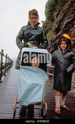 Junge kaukasische Frau im Regenmantel, die einen Kinderwagen schiebt, mit einem älteren Kind in Regenmantel, die um 1960 herumläuft. Foto archivieren Stockfoto