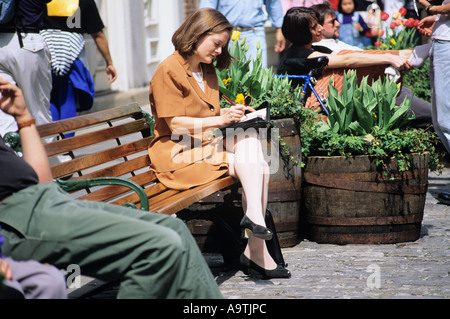 BUSINESS-FRAU SITZEN AUF BANK IN PLATZ IM FREIEN, IN BUCH SCHREIBEN Stockfoto