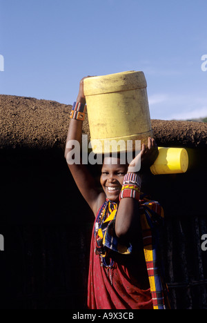 Lolgorian, Kenia. Siria Maasai Manyatta; lächelnd Maasai außerhalb einer ummauerten Lehmhaus mit einer gelben Kunststoff-Fass auf dem Kopf Stockfoto