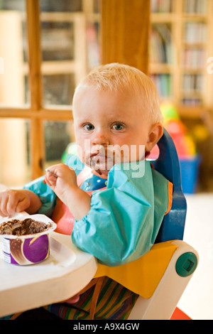 Schöne junge Baby essen Schokolade Pudding in einem Kinderstuhl in die Kamera schaut Stockfoto