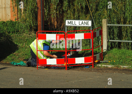 British Telecom-Techniker arbeitet an Kabeln an einem Straßen-Schach in der Grasnarbe an einem ländlichen Standort hinter einer rot-weißen Sicherheitsbarriere in einer Mill Lane Essex UK Stockfoto