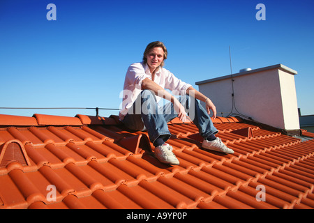 Junger Mann, der auf einem rot gekachelten Dach unter einem klaren blauen Himmel sitzt und die Aussicht und den sonnigen Tag in entspannter Pose genießt. Stockfoto