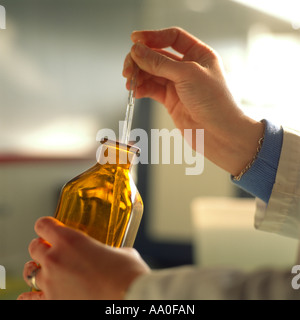 Eine Hand hält eine Pipette vorsichtig und saugt Flüssigkeit aus einer klaren, bernsteinfarbenen Flasche in einem gut beleuchteten Labor auf. Stockfoto