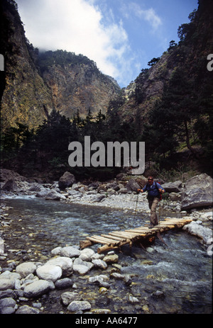 Einsamer Mann mit Rucksack überquert einen Bach durch eine provisorische Brücke mit Wanderstöcken in der sonnenbeschienenen Samaria-Schlucht, Kreta Stockfoto