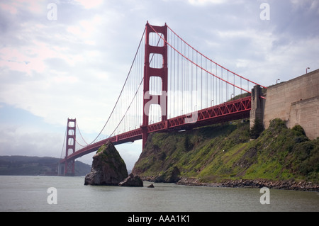 Die rote Golden Gate Bridge in San Francisco überspannt das Wasser, aus einem niedrigen Winkel nahe einer felsigen Küste unter einem bewölkten Himmel. Stockfoto