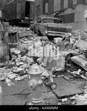 1960ER JAHRE MÄDCHEN STEHEN AUF RÜCKSEITE DREIRAD HINTER OFFENEN HYDRANTEN UMGEBEN VON TRÜMMERN MIT ALTEN KLAPPRIGEN AUTO IM HINTERGRUND Stockfoto