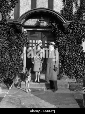 1920ER JAHREN PAAR MANTEL HUT HANDSCHUHE AUF SCHRITTE EFEU BEDECKT GEBÄUDE MIT DEUTSCHER SCHÄFERHUND AN DER LEINE Stockfoto